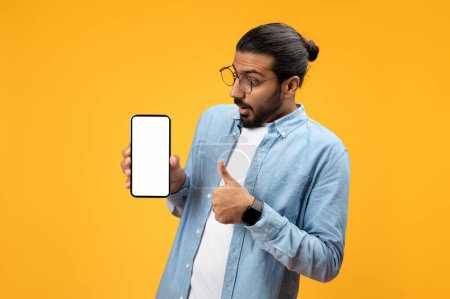 Photo for Surprised man in blue denim shirt holds up a smartphone with a blank screen and points at it with his other hand, indicating an unexpected or important notification, against a yellow background - Royalty Free Image