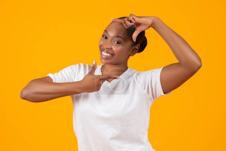 Téléchargez les photos : Joyeux jeune femme afro-américaine en t-shirt blanc fait signe cadre ou lentille avec les mains, faisant semblant de faire un geste photo créatif, sur fond de studio orange. Photographie et concept de blog - en image libre de droit