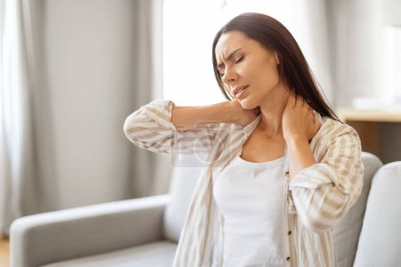 Photo for Young woman at home touching her neck with pained expression, exhausted millennial female massaging painful area and frowning, suffering osteoporosis or arthritis, sitting on couch, copy space - Royalty Free Image