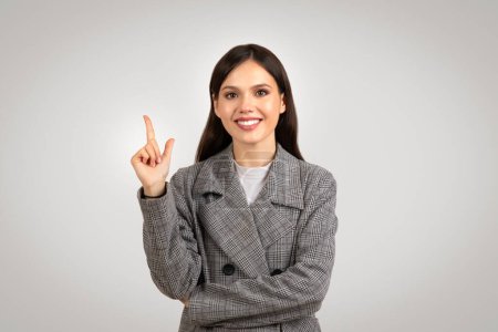 Téléchargez les photos : Une jeune femme d'affaires optimiste dans un blazer à carreaux pointe vers le haut avec un sourire éclatant, suggérant une idée créative ou une solution sur fond gris - en image libre de droit