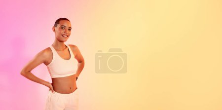 Téléchargez les photos : Une femme rayonnante avec un sourire rayonnant pose en toute confiance, les mains sur les hanches dans une tenue de fitness légère, contre un doux dégradé de teintes roses et jaunes, représentant la santé et la vitalité - en image libre de droit