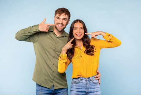 Photo for Bright and cheerful young couple with beaming smiles pointing at their white teeth, dressed in trendy green and yellow, against light blue background - Royalty Free Image