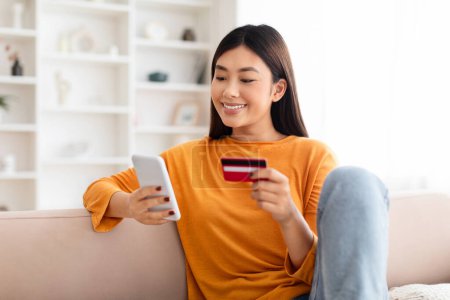 Photo for Fast Online Shopping. Smiling young chinese lady holding debit credit bank card and using cell phone, sitting on couch at home in living room, looking at smartphone screen, free copy space - Royalty Free Image