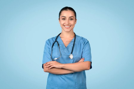 Photo for Confident and smiling female nurse in blue scrubs, arms crossed, with stethoscope around her neck, representing healthcare professionalism, blue background - Royalty Free Image