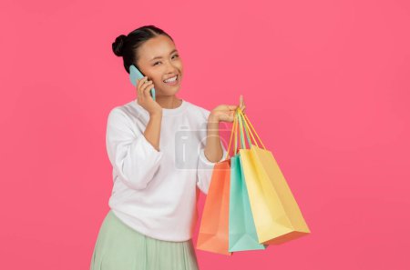 Foto de Retrato de una mujer asiática sonriente hablando por teléfono celular y sosteniendo bolsas de compras, alegre dama coreana adicta a las compras llamando a su amigo, compartiendo noticias de ventas, de pie sobre fondo de estudio rosa, espacio para copiar - Imagen libre de derechos