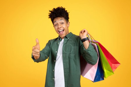 Photo for Happy emotional young black man going shopping, holding colorful paper bags and showing thumb up, standing in casual outfit over yellow studio background. Shopping, retail concept - Royalty Free Image