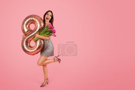Téléchargez les photos : Femme joyeuse dans une robe argentée chatoyante tient exubéramment un ballon numéro huit en or rose et un bouquet de tulipes roses sur un fond rose, célébrant une occasion spéciale - en image libre de droit