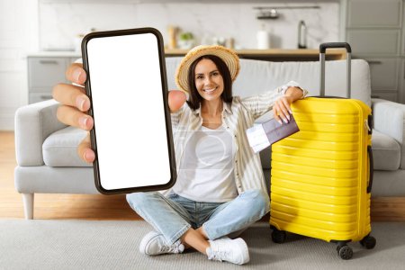 Foto de Joven turista alegre con sombrero de paja que muestra el teléfono inteligente en blanco en la cámara mientras está sentado en el suelo por el equipaje amarillo en casa, dama sonriente recomendando oferta de viaje caliente, maqueta - Imagen libre de derechos