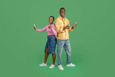 Photo for Happy millennial black man and woman with phones in their hands showing thumb up, chatting with friends or texting one another, green background. Communication, virtual world concept - Royalty Free Image