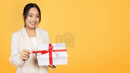 Foto de Encantada dama coreana milenaria en traje cinta de caja abierta con regalo, aislada sobre fondo de estudio amarillo, panorama. Regalo de vacaciones, cumpleaños de celebración, aniversario de felicitación - Imagen libre de derechos