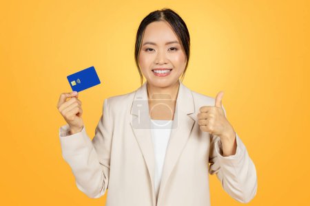 Téléchargez les photos : Femme d'affaires japonaise millénaire souriante en costume, montrer carte de crédit et pouce vers le haut, isolé sur fond orange. Recommandation de finances, argent pour payer et acheter, vendre et faire du shopping - en image libre de droit