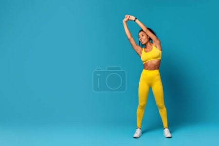 Athletic Black Woman In Yellow Fitwear Doing Core Side Bend With Raised Arms, While Stretching And Warming Up Body During Workout Against Blue Studio Background. Full Length Shot, Copy Space