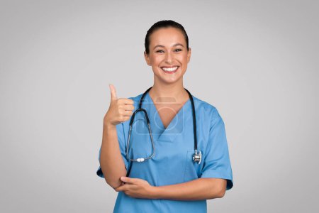 Téléchargez les photos : Infirmière confiante et souriante en blouse bleue donnant son signe du pouce vers le haut, suggérant l'approbation ou le succès dans un cadre de soins de santé, avec un fond gris - en image libre de droit