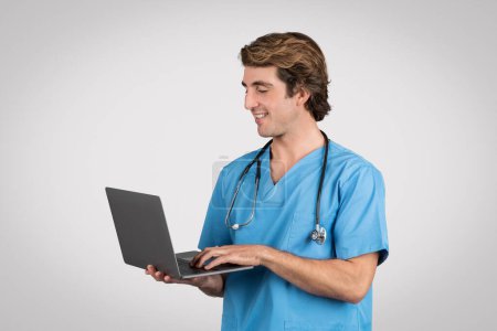 Téléchargez les photos : Infirmière engagée en blouse bleue utilisant joyeusement un ordinateur portable, représentant l'intégration des technologies modernes dans les soins de santé, sur un fond gris - en image libre de droit