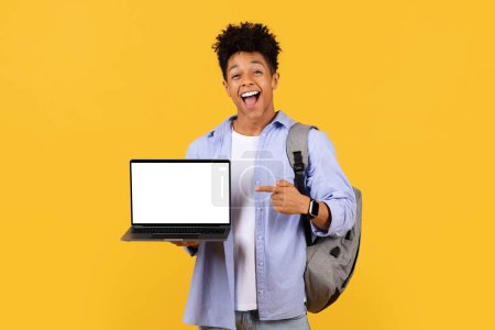 Foto de Estudiante masculino joven exuberante que presenta la pantalla en blanco del ordenador portátil, perfecta para el espacio de la copia, con la expresión alegre, fijada contra fondo amarillo soleado, maqueta - Imagen libre de derechos