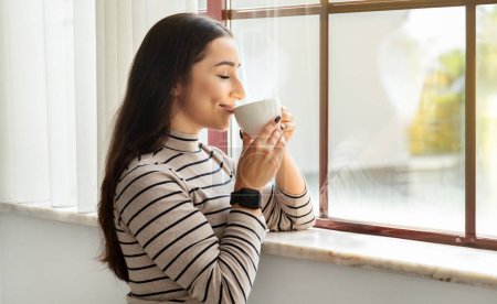 Photo for Contented young woman with sleek hair in a smartwatch and striped shirt savors her coffee by a sunlit window, embodying a relaxed lifestyle. Coffee break, morning routine drink - Royalty Free Image