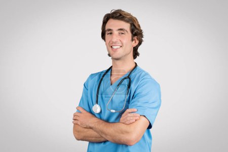 Happy male nurse wearing blue scrubs and stethoscope with arms confidently folded, embodying professional medical care, against light gray background