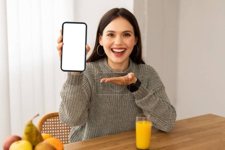 Photo for Nutrition mobile app, diet, detox, healthy lifestyle and gadgets. Cheerful young woman showing phone with white blank screen, drinking fresh orange juice, eating organic fruits, home interior - Royalty Free Image