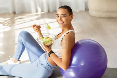 Téléchargez les photos : Joyeux sourire femme latine assis sur un ballon d'exercice à la maison, manger une salade fraîche, illustrant un mode de vie sain qui combine l'activité physique avec des aliments nutritifs - en image libre de droit