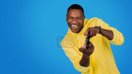 Foto de Un hombre negro encantado con una sudadera con capucha amarilla juega alegremente con un controlador de juego, su expresión llena de emoción y diversión contra un impresionante fondo de estudio azul, panorama - Imagen libre de derechos