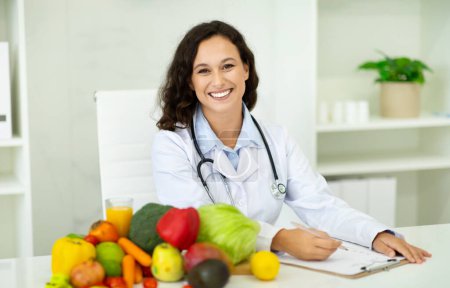 Photo for Choose quality healthy food. Happy european woman nutritionist sit at desk with fresh fruits and vegetables, smiling at camera at clinic. Weight loss consultant recommending healthy plant based diet - Royalty Free Image