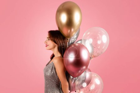 Foto de Una mujer serena con un vestido plateado brillante mira a lo lejos, su perfil acentuado por un montón de elegantes globos metálicos sobre un cálido fondo rosa. Evento de celebración - Imagen libre de derechos