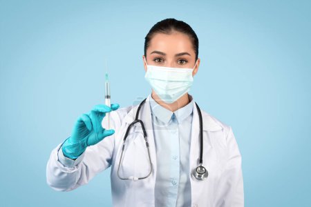 Ernsthafte Ärztin in OP-Maske und Handschuhen zeigt vorsichtig eine Spritze, bereit für Impfungen oder Behandlungen, blauer Hintergrund