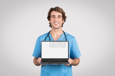 Cheerful male nurse in blue uniform holding an open laptop with white blank screen for healthcare information and records, mockup