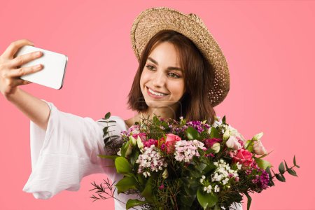 Foto de Una radiante joven europea feliz en un traje de verano elegante y sombrero de paja toma una selfie con un vibrante ramo de flores mixtas en el teléfono sobre un fondo rosa, estudio - Imagen libre de derechos