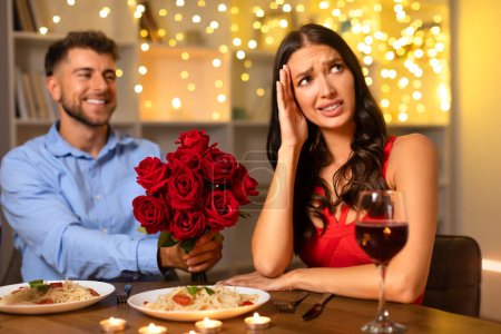 Photo for Happy man offering bouquet of roses, with stressed woman holding her head in hand at a candlelit dinner, amid festive lights - Royalty Free Image