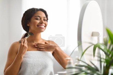 Jeune femme noire massant le cou avec un rouleau de jade en pierre verte près du miroir, souriante femme afro-américaine s'engageant dans une routine de beauté auto-soin dans le confort de sa maison, favorisant la santé de la peau