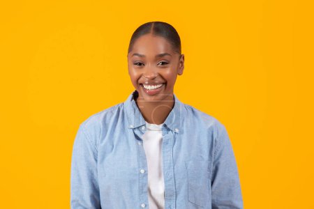 Téléchargez les photos : Portrait de jolies jeunes afro-américaines posant montrant un beau sourire, exprimant la confiance et le style décontracté moderne en chemise en denim, sur fond de studio jaune - en image libre de droit