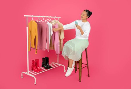 Foto de Shopaholic señora asiática sosteniendo Smartphone y mirando en rack con ropa de moda, Mujer coreana feliz usando aplicación móvil para compras en línea, Sentado en la silla sobre fondo rosa en el estudio, espacio libre - Imagen libre de derechos