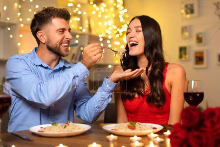 Photo for Happy couple sharing moment, with cheerful man feeding spaghetti to an amused woman, both enjoying a candlelit dinner with festive lights - Royalty Free Image