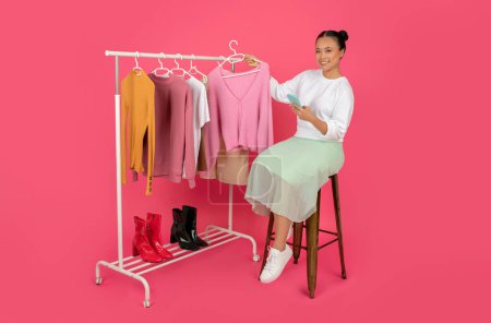 Photo for Personal Stylist App. Happy Asian Woman With Smartphone Sitting On Chair Near Clothing Rack, Smiling Korean Female Fashion Consultant Demonstrating New Clothes, Posing On Pink Background - Royalty Free Image