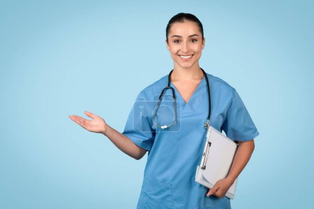 Foto de Sonriente profesional de la salud en uniformes azules sosteniendo un portapapeles y haciendo gestos con la mano para presentar información de salud o un espacio vacío - Imagen libre de derechos