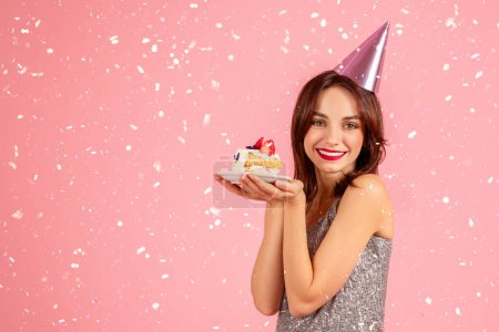 Foto de Encantada mujer caucásica milenaria con un vestido brillante y un sombrero de fiesta, sosteniendo un pedazo de pastel de cumpleaños con confeti cayendo a su alrededor, celebrando alegremente sobre un fondo rosa - Imagen libre de derechos