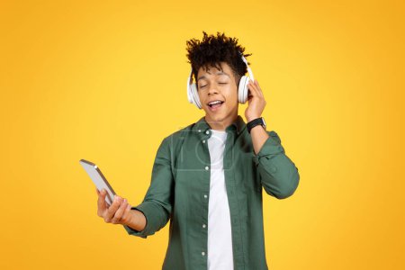Téléchargez les photos : Joyeux heureux élégant jeune noir écoute de la musique sur fond de studio jaune, en utilisant des écouteurs sans fil et smartphone, profiter de chansons préférées, copier l'espace - en image libre de droit