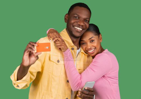 Photo for Cute young black couple with phone showing bank credit card, happy african american millennial man and woman shopping and banking online, hugging over green studio background - Royalty Free Image