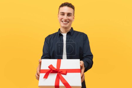 Photo for Smiling cool blonde caucasian young man giving present white gift box with red bow, celebrating anniversary or St. Valentines Day with his girlfriend, yellow studio background - Royalty Free Image