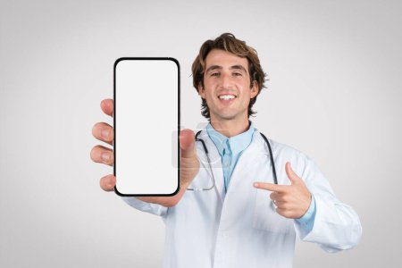Téléchargez les photos : Médecin masculin joyeux en blouse blanche pointant vers un smartphone avec écran blanc, parfait pour afficher des applications médicales ou des solutions de télésanté, une maquette - en image libre de droit