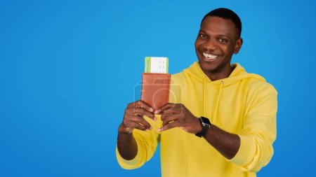 Foto de Un hombre afroamericano radiante en una sudadera con capucha amarilla mientras sostiene su pasaporte y su tarjeta de embarque, listo para una aventura, contra un audaz fondo de estudio azul, panorama - Imagen libre de derechos