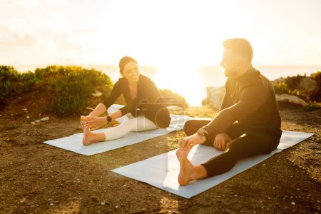 Photo for Cheerful couple enjoying their yoga routine, stretching on mats by the sea, with the golden sunlight of dusk creating warm, inviting atmosphere - Royalty Free Image