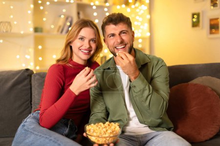 Téléchargez les photos : Couple ravi de partager rire tout en mangeant du pop-corn sur un canapé confortable, avec une toile de fond de lumières scintillantes de fées ajoutant de la chaleur à la scène - en image libre de droit