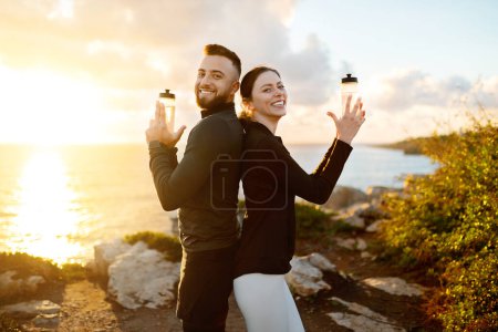 Foto de Hombre y mujer sonrientes espalda con espalda, sosteniendo alegremente botellas de agua como si fueran armas, durante la puesta de sol radiante al aire libre en la orilla del mar - Imagen libre de derechos