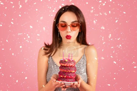 Photo for Happy millennial caucasian woman in dress, glasses blows out the candles makes a wish with cake donuts, isolated on pink background, studio. Celebration holiday, birthday party with confetti - Royalty Free Image