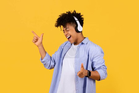 Foto de Alegre estudiante adolescente negro con auriculares perdidos en la música, señalando y cantando, atrapado en el momento de la dicha en el fondo amarillo soleado - Imagen libre de derechos