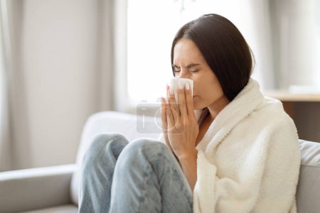 Foto de Retrato de una mujer joven enferma envuelta en una manta estornudando la nariz en el tejido, una mujer milenaria enferma que sufre de resfriado o gripe, sentada en el sofá en la sala de estar brillante en casa, espacio para copiar - Imagen libre de derechos