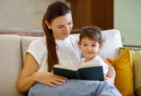 Photo for Loving young mother and her cute little daughter reading book and embracing, cute preschool girl and mom relaxing on couch in living room, bonding together, enjoying spending time with each other - Royalty Free Image