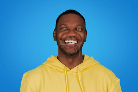 Téléchargez les photos : Un homme afro-américain heureux dans un sweat à capuche jaune rayonne avec un sourire radieux, mettant en valeur les dents blanches sur un fond bleu brillant, exsudant bonheur et positivité - en image libre de droit
